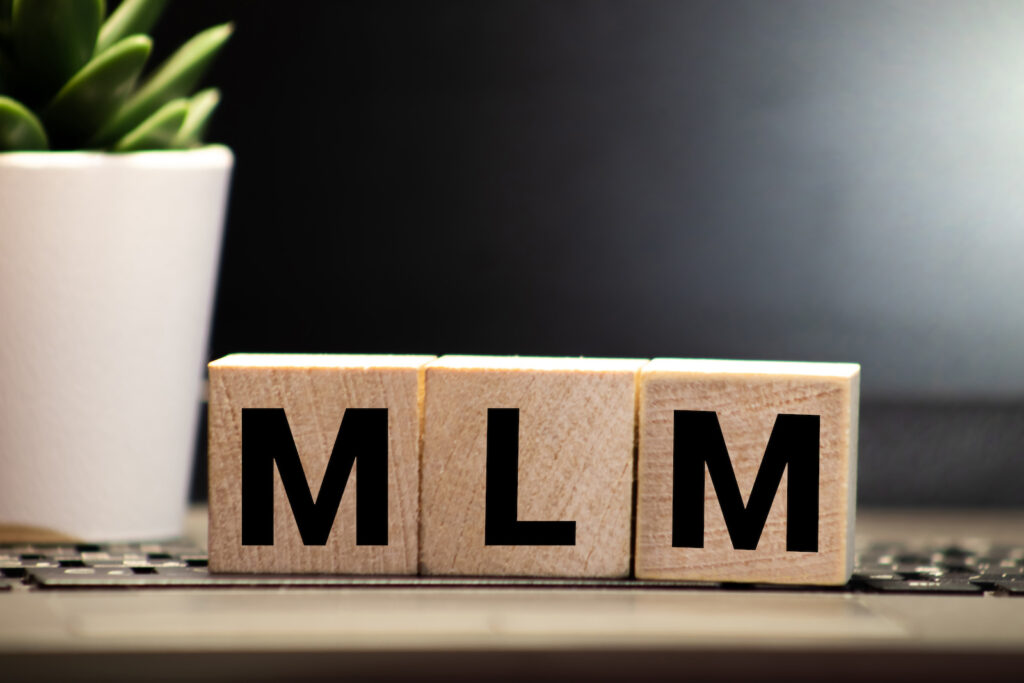 Image of MLM Multi-Level Marketing written on a wooden cube in a office desk.